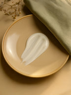 Beige plate with a dollop of white cream on a beige surface with green fabric and flowers. White conditioner dollop on a warm beige ceramic plate with a sage linen cloth, lemon slice, and subtle lavender sprig under soft natural daylight—showing glossy, creamy texture.