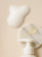 Marble-textured bottle with pump and droplet on beige background Foaming pump of Sensitive Skin Face & Body Cleanser at bottom; round marble coaster above with gel droplet and airy foam on beige linen.