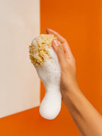 Hand holding a white scrubber with a natural fiber head against an orange and beige background Close-up of creamy white Suvéra Hand & Body Wash Grapefruit foam with soft bubbles and a hint of peach tone, captured in warm natural light for a fresh and luxurious self-care feel.