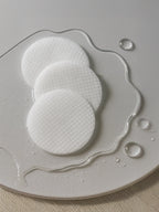 Three white round cotton pads on a gray plate with water droplets. Hydrating Toner