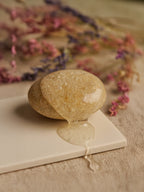Stone with water droplets on a white surface with pink flowers in the background Suvéra Nourish & Volumize Shampoo — amber gel streaming over a smooth river pebble on linen, dried flowers softly blurred, natural daylight, self-care hair care.