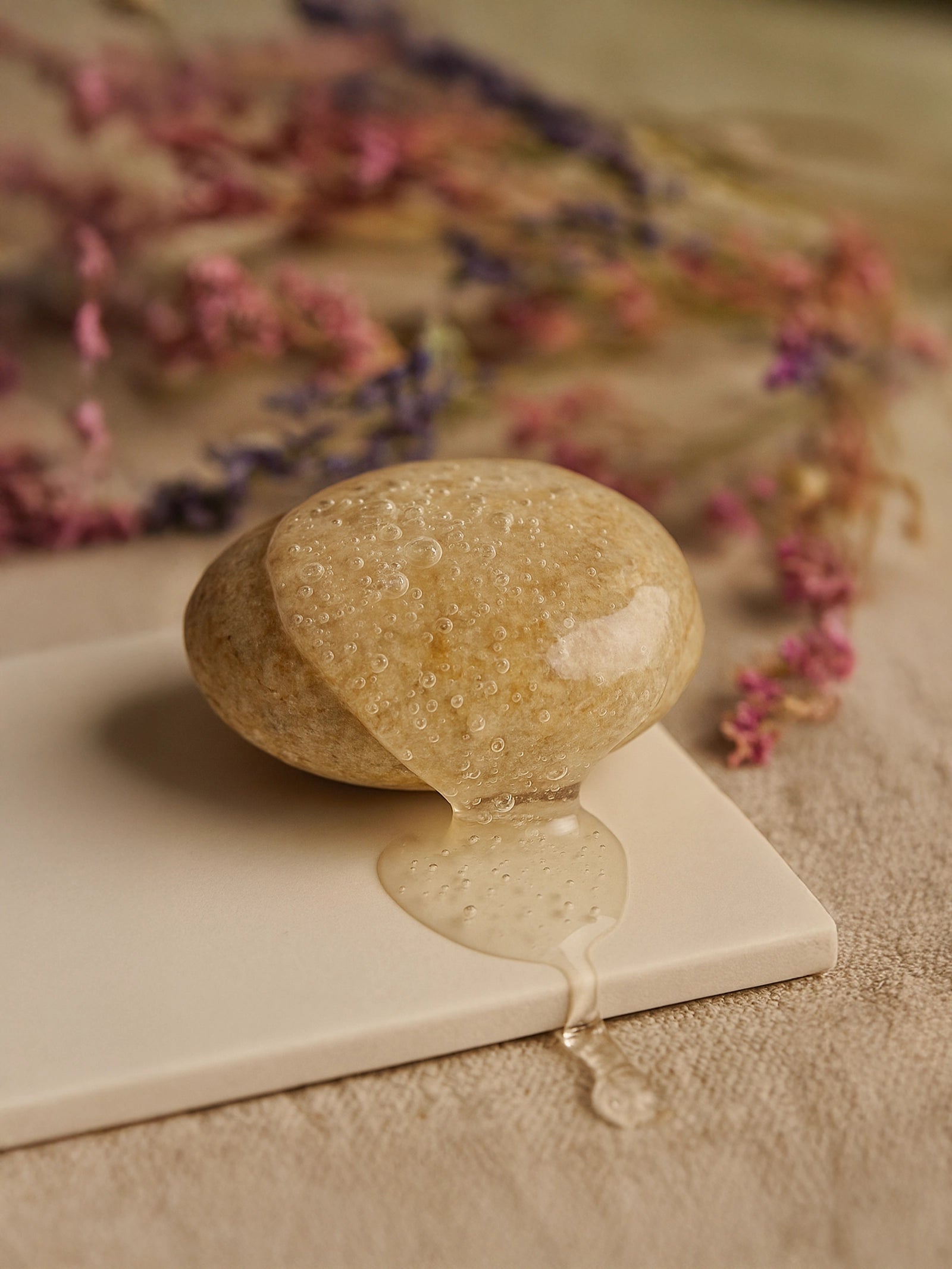 Stone with water droplets on a white surface with pink flowers in the background Suvéra Nourish & Volumize Shampoo — amber gel streaming over a smooth river pebble on linen, dried flowers softly blurred, natural daylight, self-care hair care.
