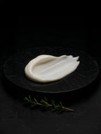 Front-facing smear of white body cream on dark marble slab, overhead light; fresh thyme sprigs in the foreground.