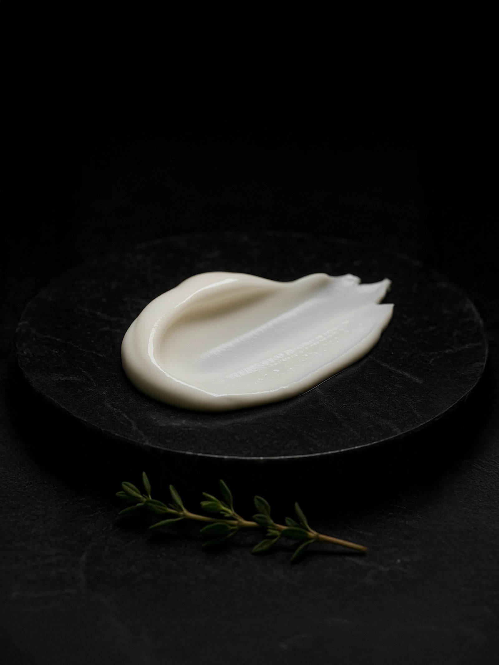 Front-facing smear of white body cream on dark marble slab, overhead light; fresh thyme sprigs in the foreground.