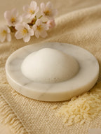 White round bath bomb on a marble soap dish with flowers and rice grains on a beige fabric background